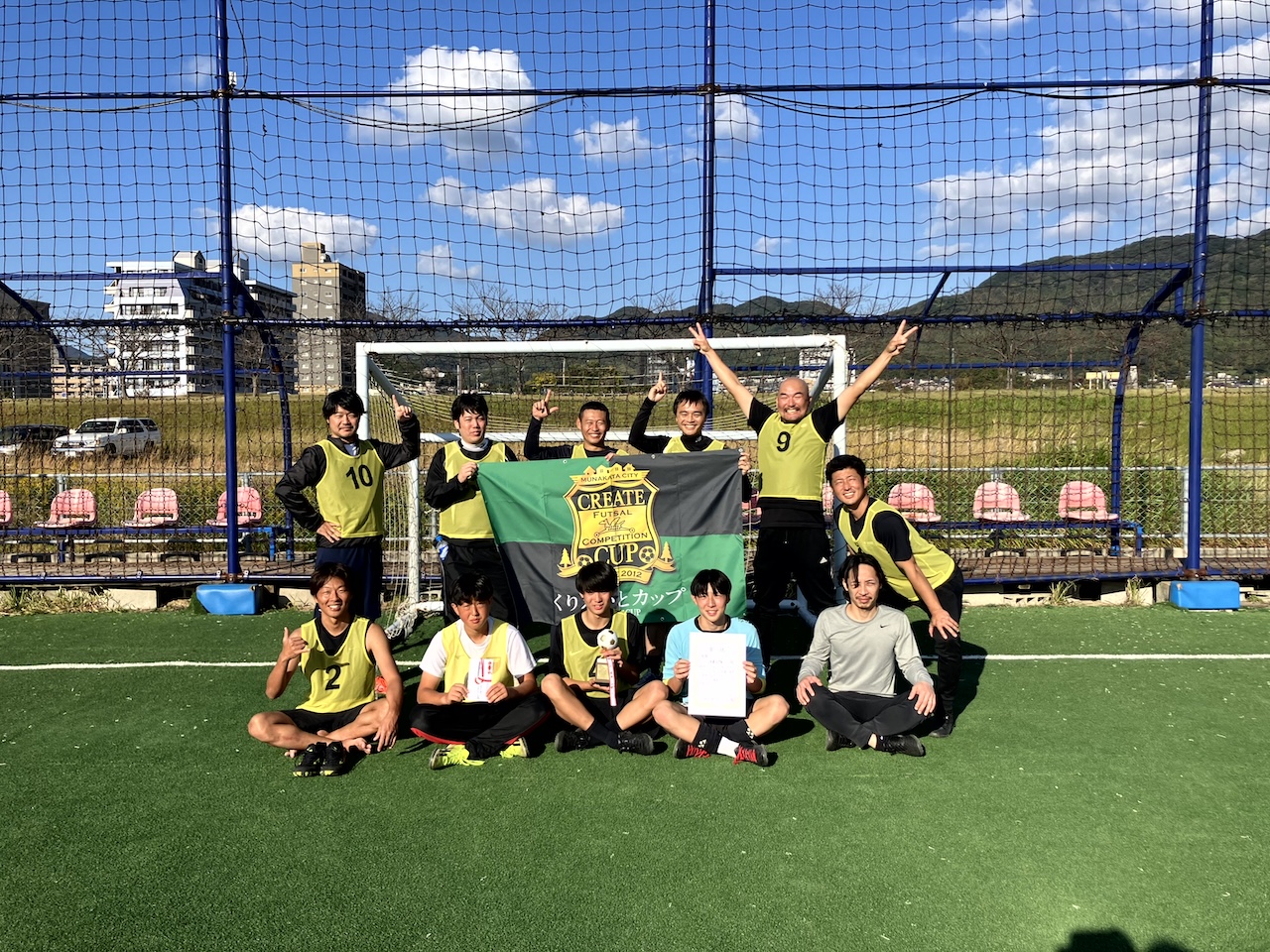 仲間とつかんだ栄冠 河東OYGが優勝旗とともにフィールドで記念撮影　JOY FIELD 15 MUNAKATA 第12回くりえいとカップ 優勝チーム 河東OYG