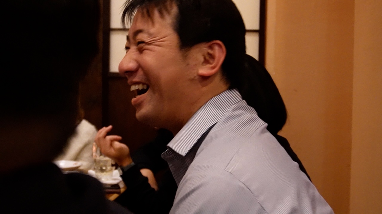 飲食店の席で大きく笑っている男性。リラックスした雰囲気で、周囲と楽しく会話している様子