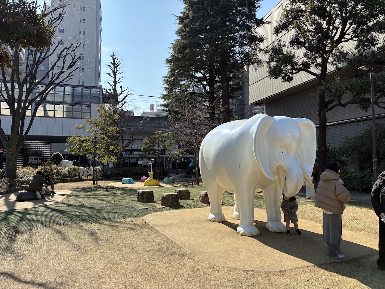 都市部の公園に大きな白い象のオブジェがあり、子どもたちや親子連れが遊んでいる様子