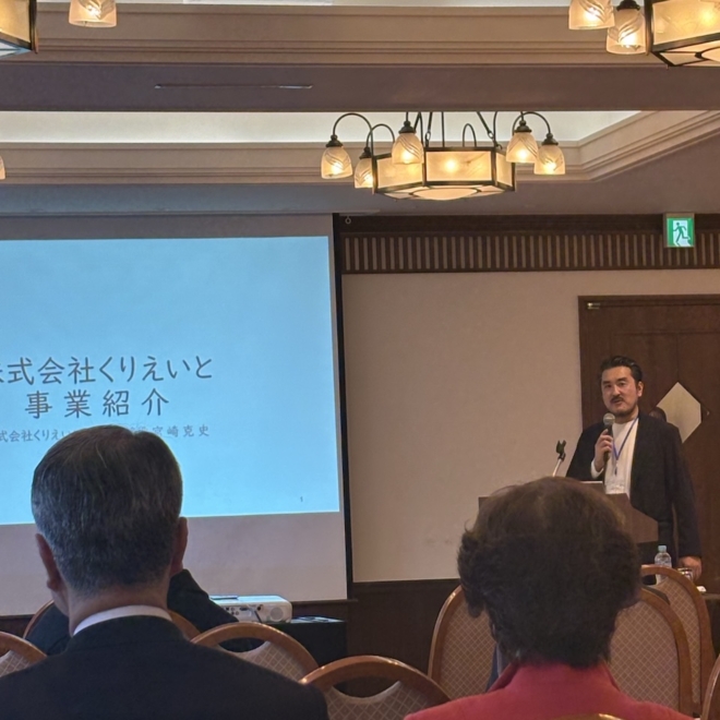 会場でマイクを持ち、株式会社くりえいとの事業紹介スライドの前で登壇する様子