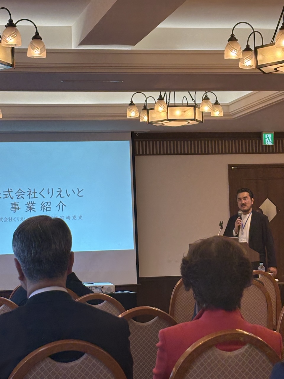 会場でマイクを持ち、株式会社くりえいとの事業紹介スライドの前で登壇する様子