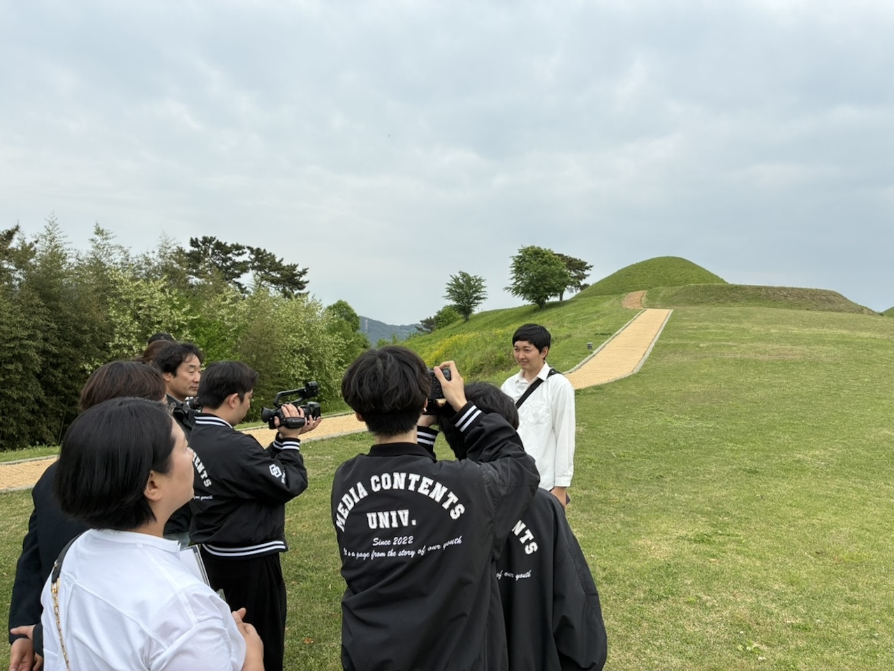 韓国・金海市近郊の古墳エリアでマサン大学の学生から映像取材を受ける宗像経済新聞の中村さん