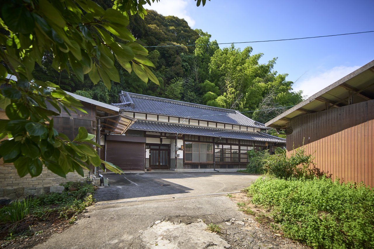 緑の山を背景に佇む古民家の母屋と納屋、引き込み道から見た敷地全景記事で「枡形」と表現した引き込み道からの眺め。農機具小屋が視界を遮る配置がわかる