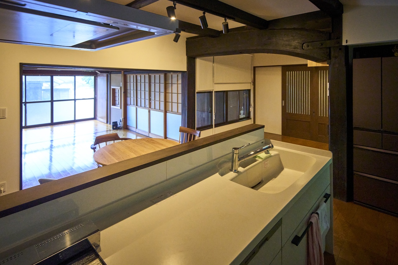対面キッチンから見た古民家の共用部。太い黒梁と障子越しにダイニングテーブルの並ぶリビングが見える