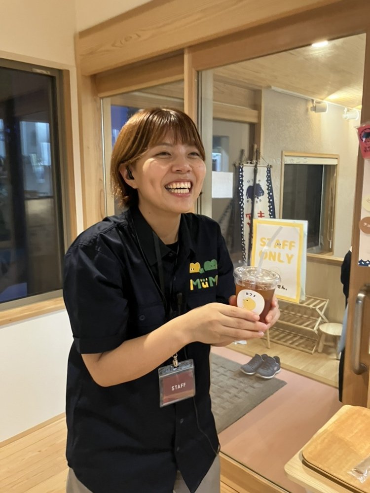 Mu-Moスタッフシャツを着た女性がアイスドリンクを手に笑顔で立っている様子