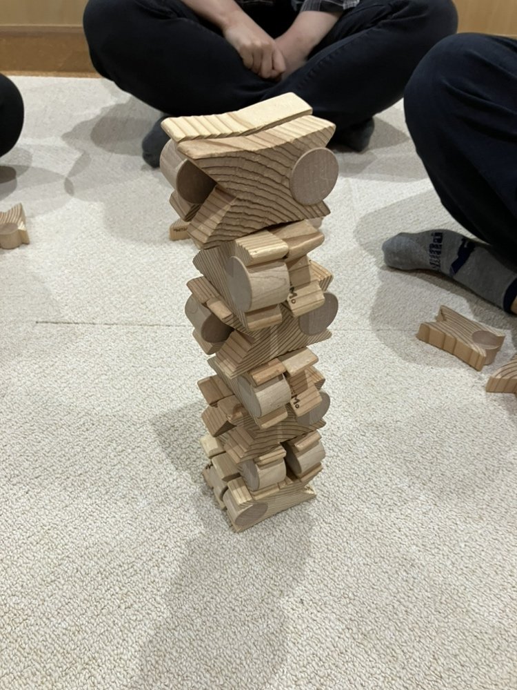 床の上に高く積み上げられた木製つみぼぼタワーと見守る参加者の手元