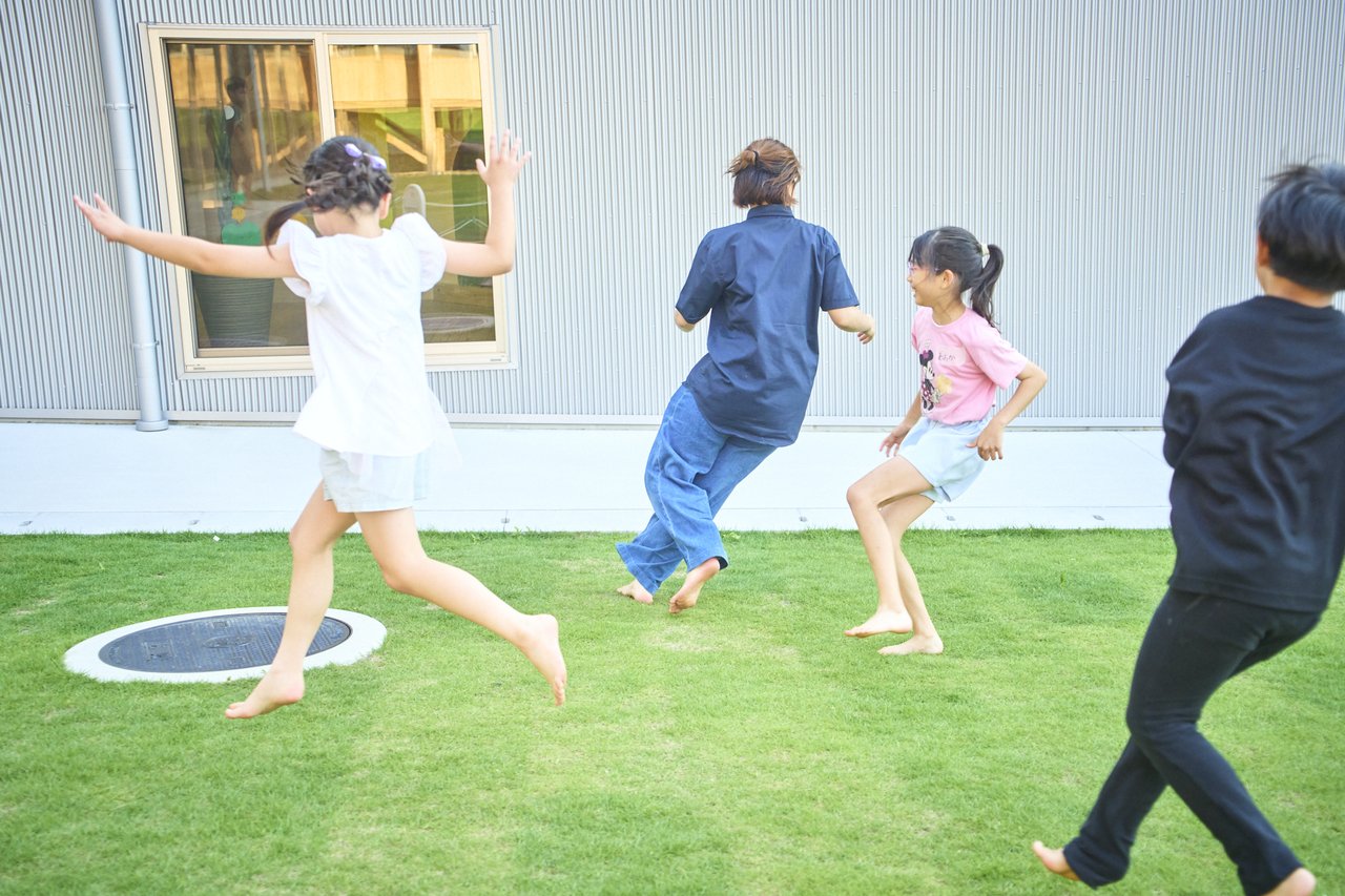 芝生の上を裸足で駆け回るスタッフと子どもたちの後ろ姿