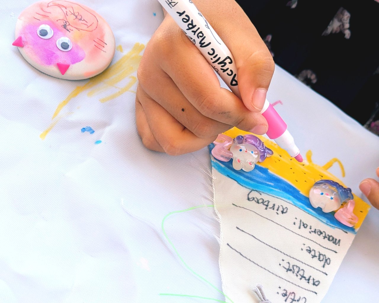 子どもの手がアクリルマーカーで布製タグにカラフルな絵を描いている