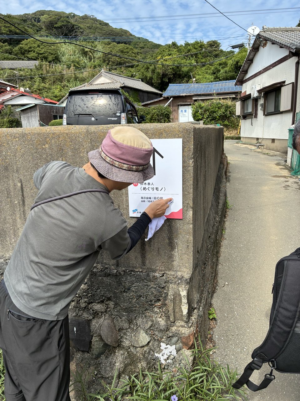 帽子をかぶった男性が島の路地でアート展示の案内看板を設置している