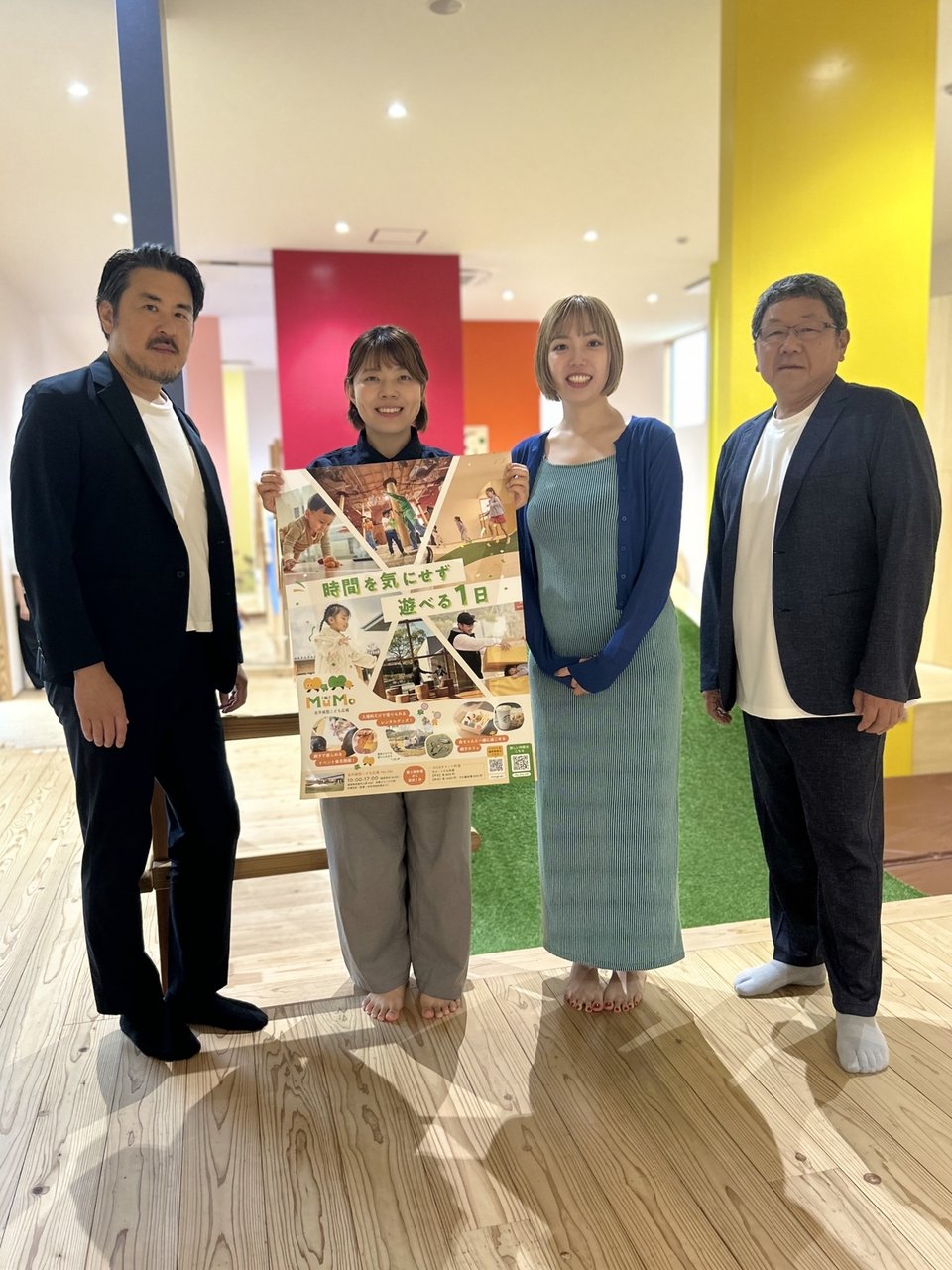 Mu-Moのポスターを持つ4名のスタッフがカラフルな室内で記念撮影している様子