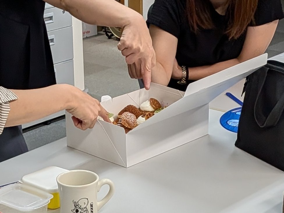 オフィスでスタッフが白い箱に入ったシュークリームを指差しながら選んでいる様子
