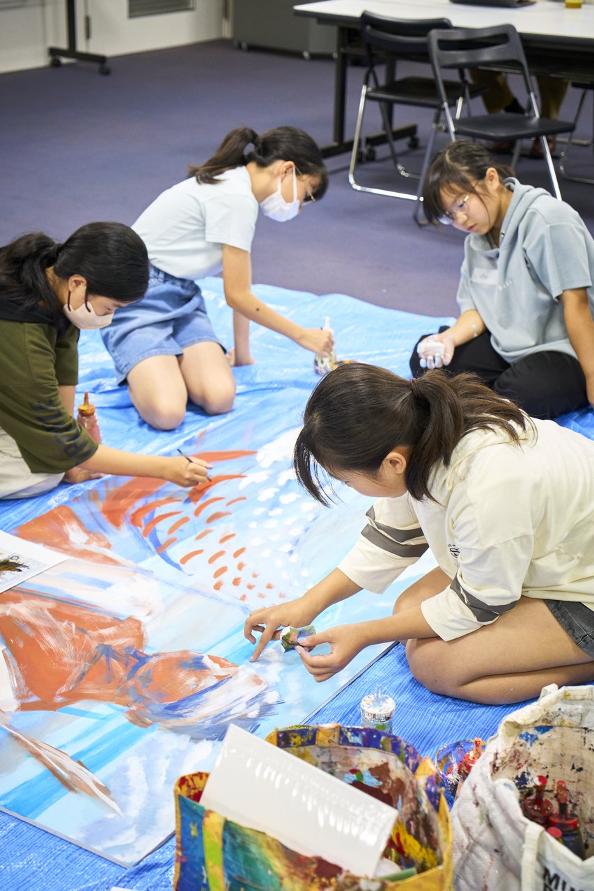 中学生たちがブルーシートの上で大きなキャンバスに絵を描いている制作風景