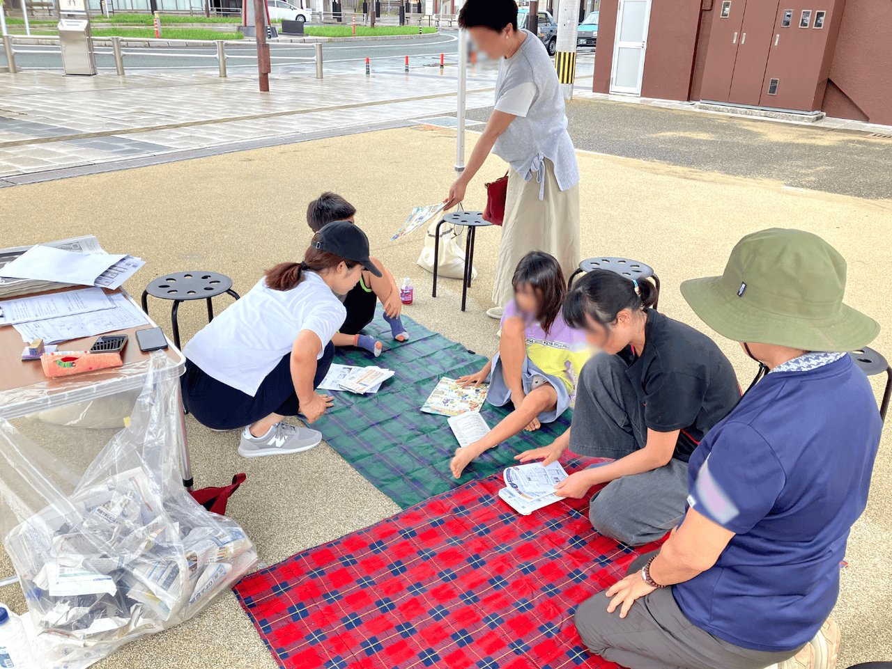 レジャーシートの上で新聞紙を使った防災グッズづくりに取り組む親子とスタッフ