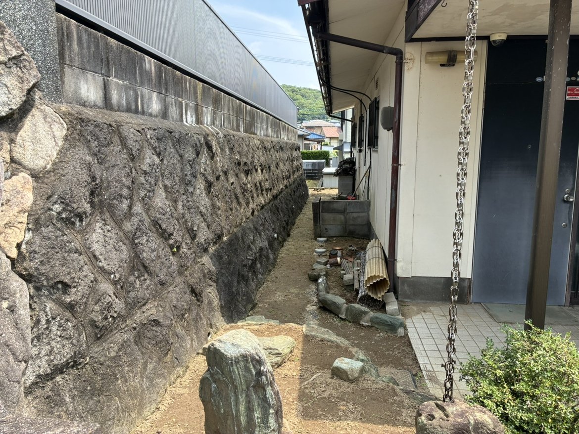 建物脇の通路と隣地境界の石積み擁壁、雨樋チェーンが見える現況